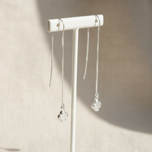 A pair of silver dangle earrings with clear faceted crystal drops, displayed on a white jewelry stand against a warm beige textured background.