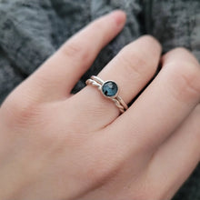 Hand wearing a silver ring with a teal kyanite gemstone against a textured gray background